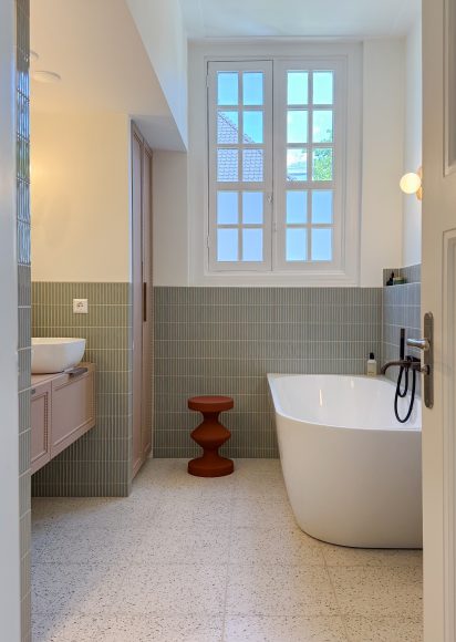 Moderne badkamer met vrijstaand wit bad onder dubbele openslaande ramen, groene tegelmozaïek wandbekleding tot halve hoogte, terrazzo vloer en zwevend badkamermeubel. Oranje designkrukje als accent en ronde wandlamp voor sfeerverlichting.