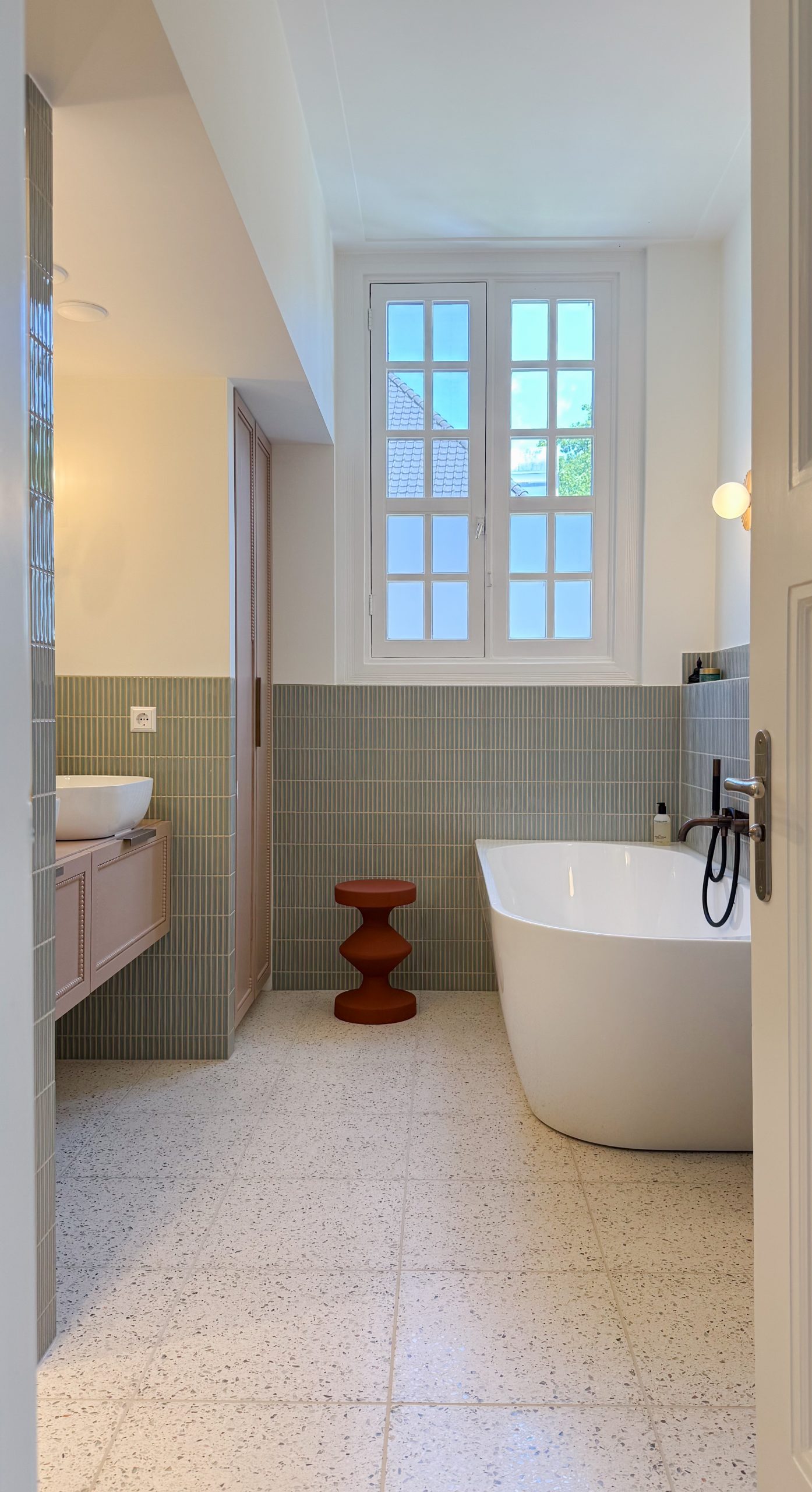 Moderne badkamer met vrijstaand wit bad onder dubbele openslaande ramen, groene tegelmozaïek wandbekleding tot halve hoogte, terrazzo vloer en zwevend badkamermeubel. Oranje designkrukje als accent en ronde wandlamp voor sfeerverlichting.