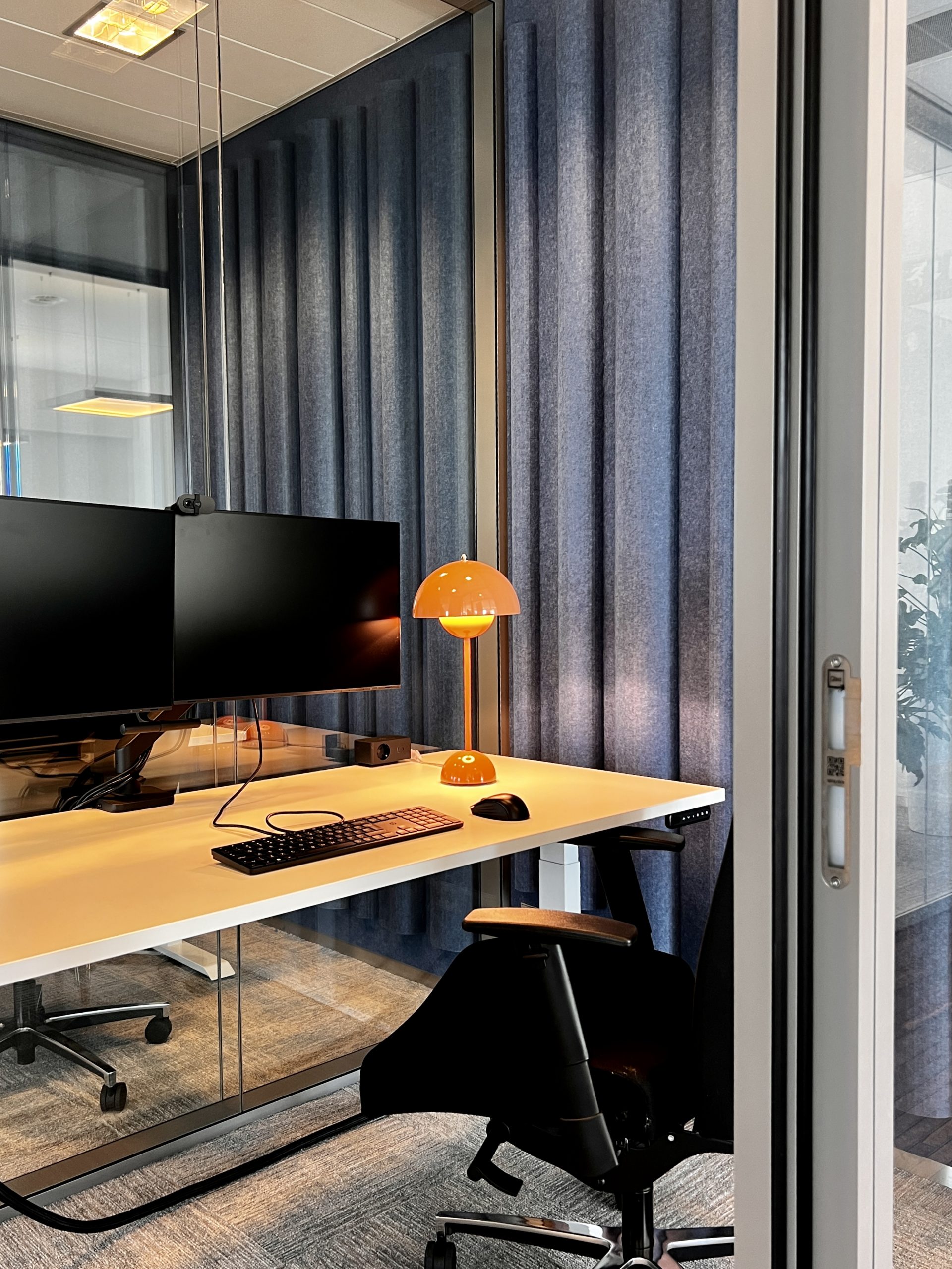 Modern kantoor met glazen werkruimte, wit bureau en zwarte ergonomische stoel. Opvallende roze designlamp op het bureau en blauwe akoestische panelen als wandbekleding. Zwarte monitor en toetsenbord completeren de werkplek.