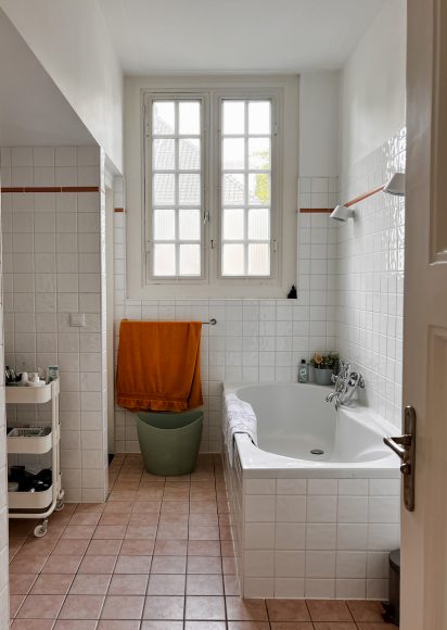 Badkamer in originele staat voor de renovatie. Witte tegels met oranje rand, ingebouwd bad met dubbele openslaande ramen erboven, groene emmer, oranje handdoek en trolley met badproducten. Bruine tegelvloer en witte wanden tonen de uitgangssituatie.
